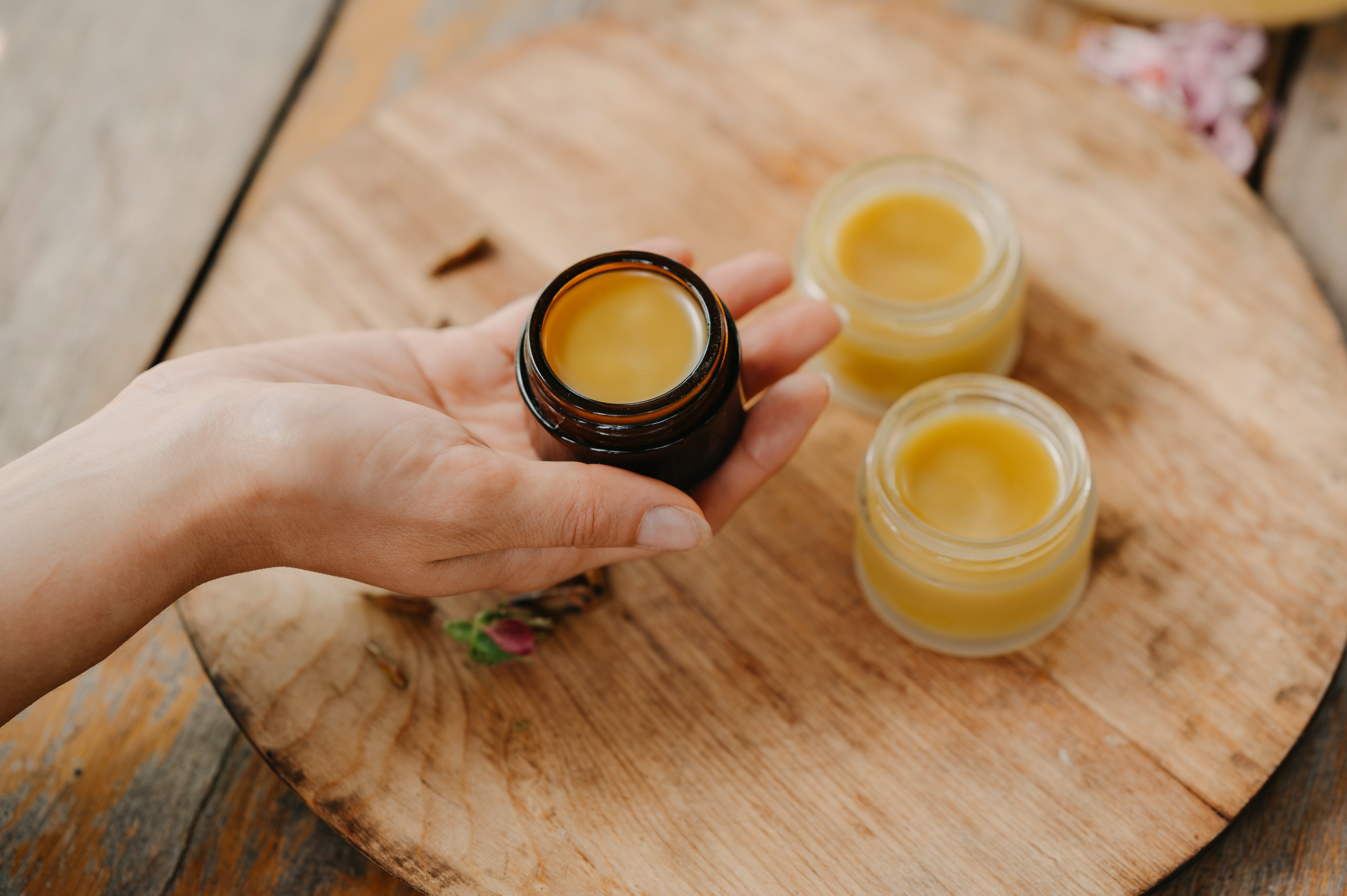 Close-up of hand presenting open jar filled with yellow cream, with two matching jars on rustic wooden board, warm light, handmade skincare product styling, natural beauty concept