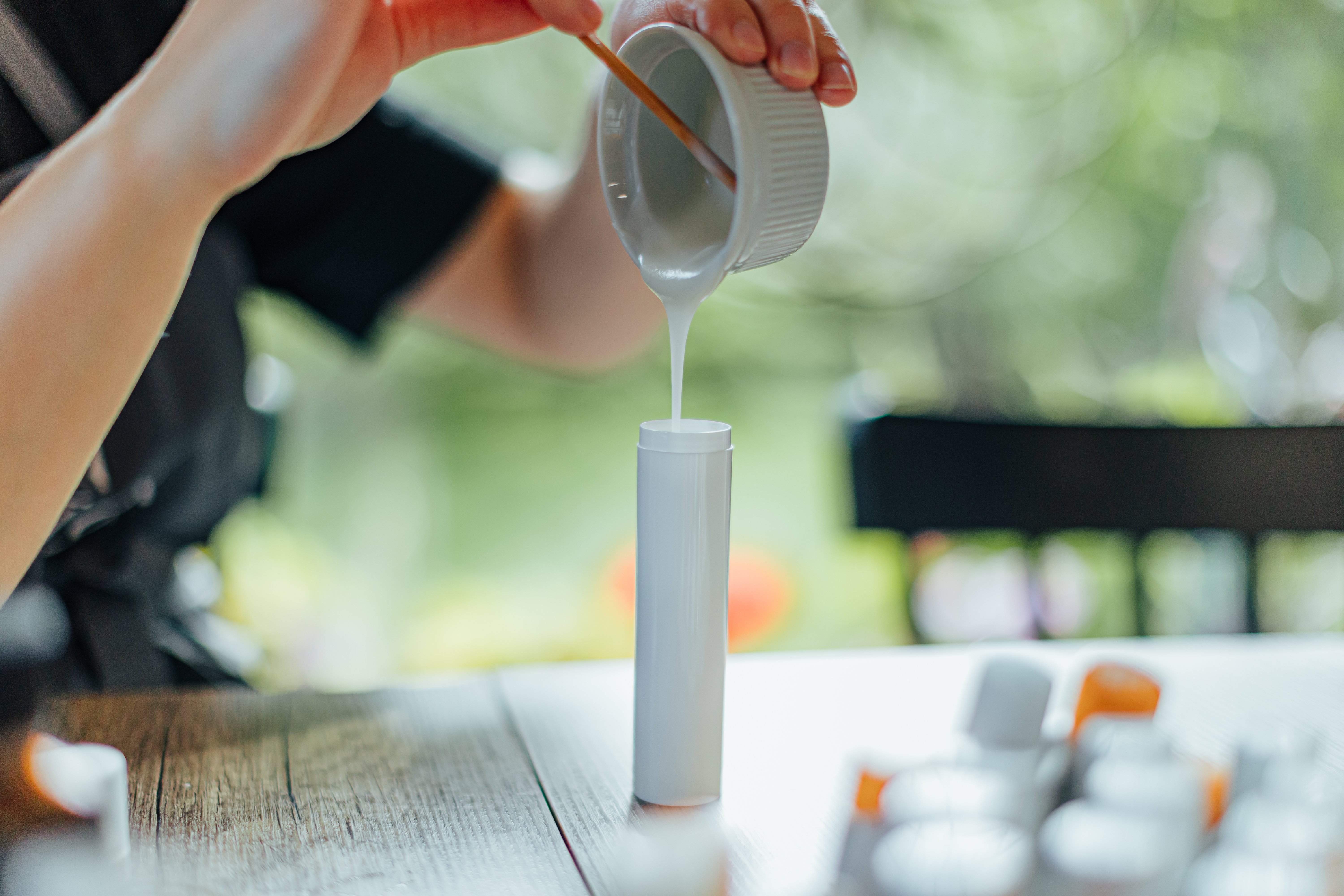 Hands pour a smooth white cream from a ceramic ramekin into a tall unbranded cosmetic tube, with blurred containers and soft green bokeh in the background